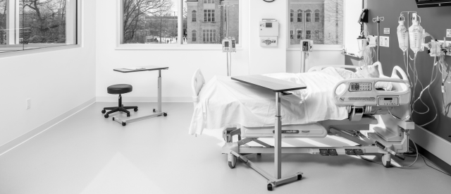 一个现代人的单色照片, 空的医院房间里有一张床, 各种医疗设备, 还有一扇可以俯瞰外面建筑的大窗户.